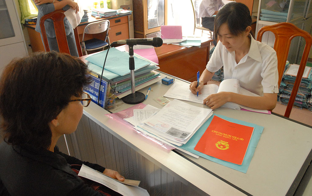 hai người đang trao đổi về nhiều giấy tờ trên bàn, bên cạnh là cuốn sổ đỏ