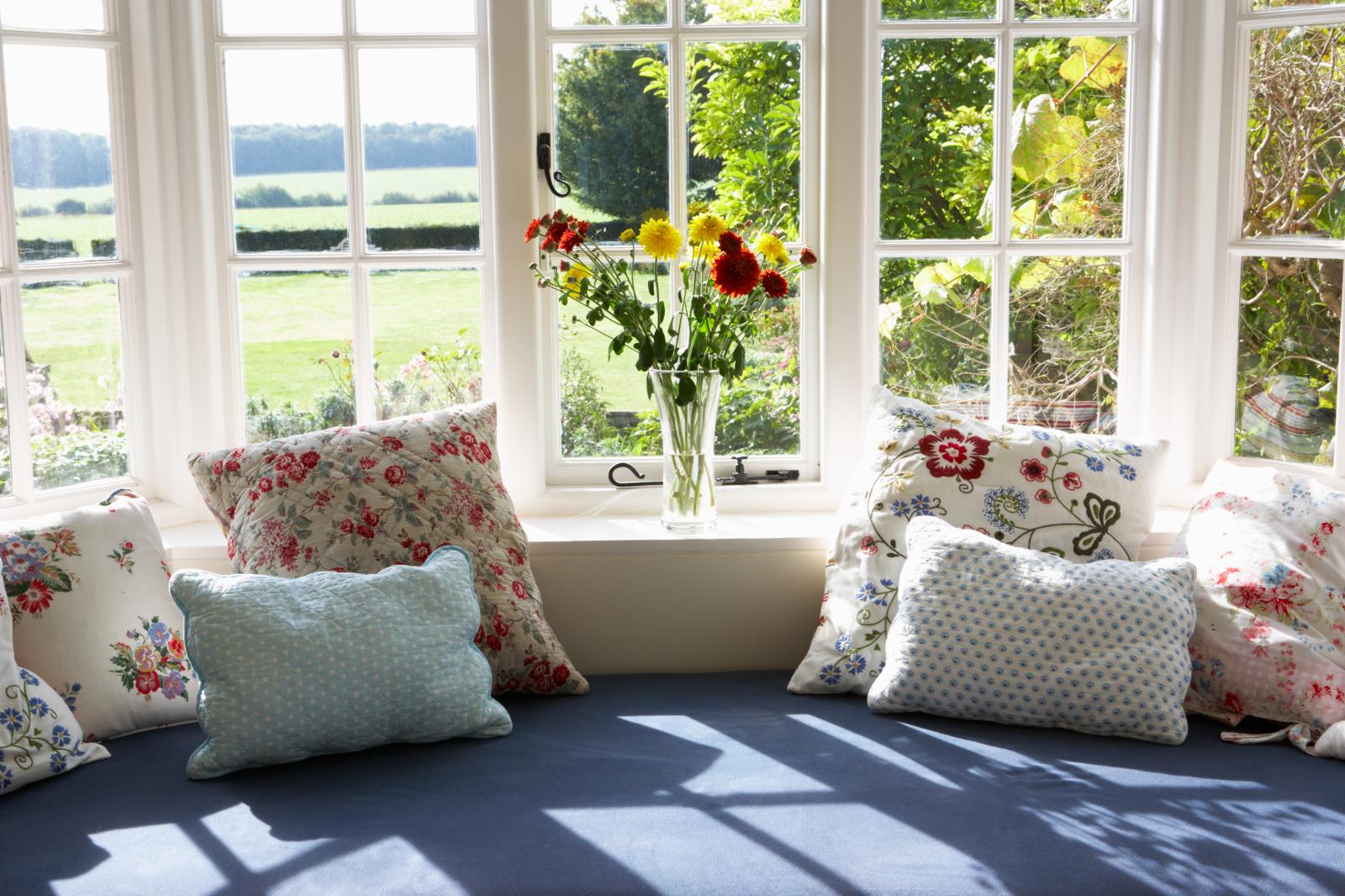 Ghế sofa có nhiều gối ôm đặt cạnh khung cửa sổ kính trắng nhìn ra bãi cõ, lọ hoa nhiều màu.