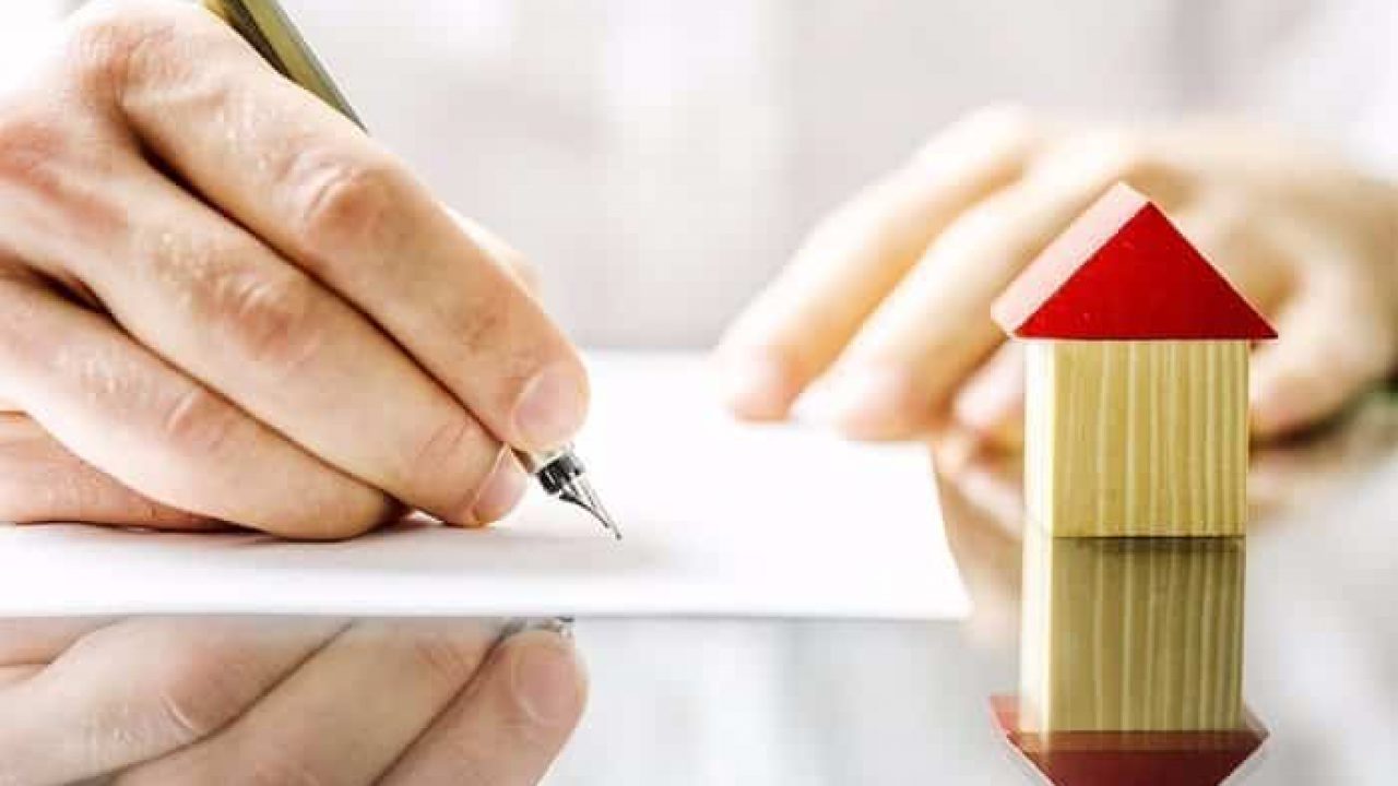 một bàn tay cầm bút viết lên tờ giấy, bên cạnh là mô hình ngôi nhà